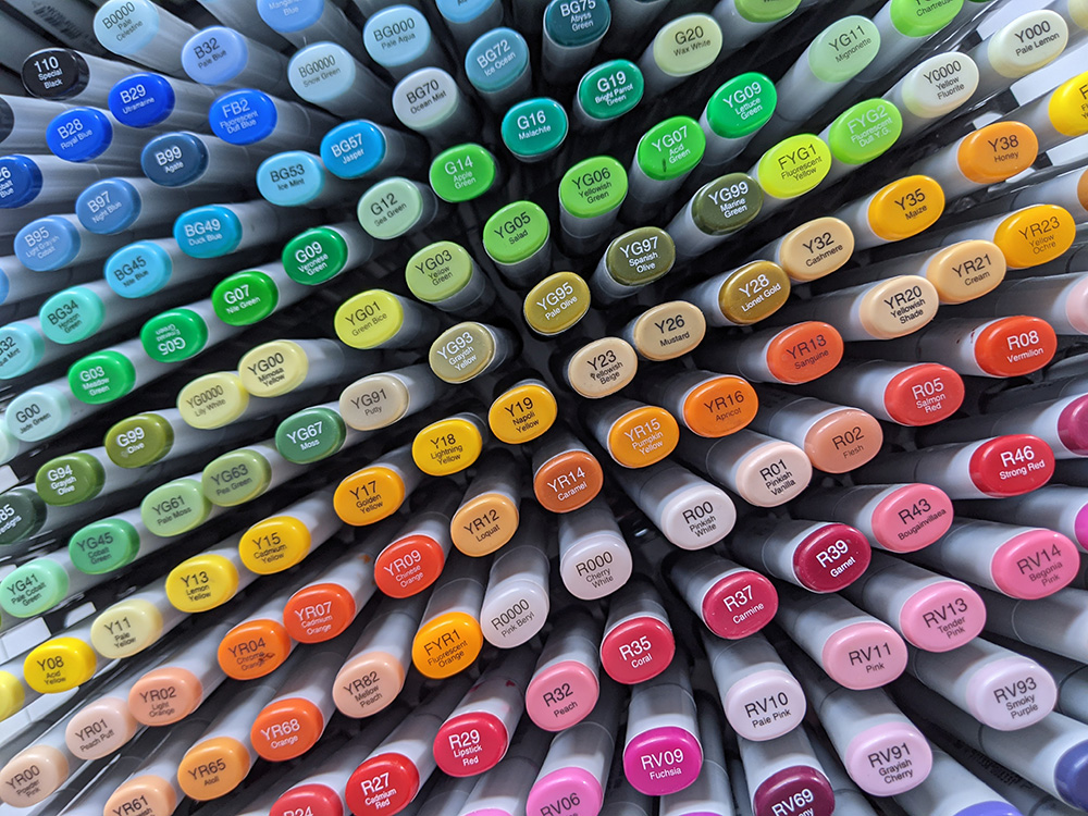 Collection de feutres japonais de la marque Copic. L'image est composée d'une vue du dessus de tous les feutres rangés verticalement. Les bouchons sont colorés, portent la numérotation de chaque feutre. L'ensemble forme un bel arc-en-ciel.