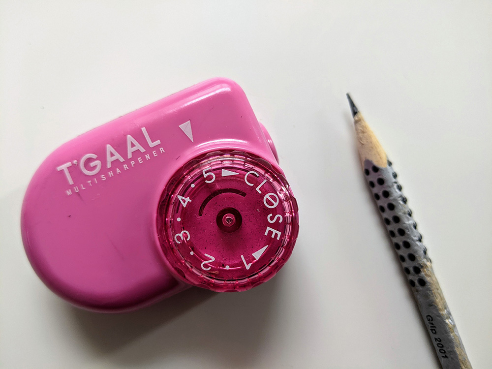 Taille crayon en plastique rose, T'Gaal écrit sur l'objet. Une molette est graduée de 1 à 5. Un crayon graphite Faber Castell à la mine très bien taillée est posé à côté.